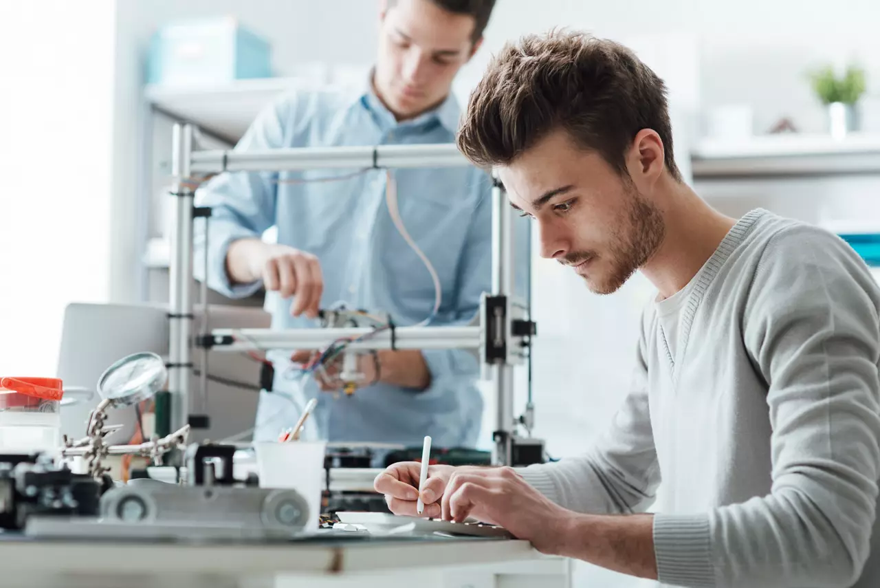 Two male engineering students working in the lab, a student is using a 3D printer in the background