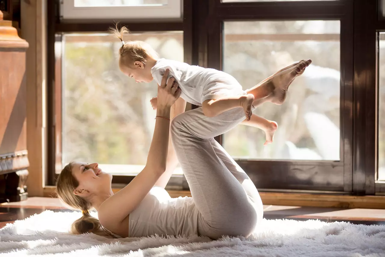 Young mother working out wearing white sportswear,  exercising at home with baby daughter, leg lifting with kid as a weigh, exercising and bonding with child, enjoyment. Healthy lifestyle concept