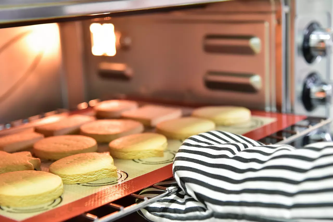 Close up hand in oven mitt taking cookies out of the oven in the kitchen.