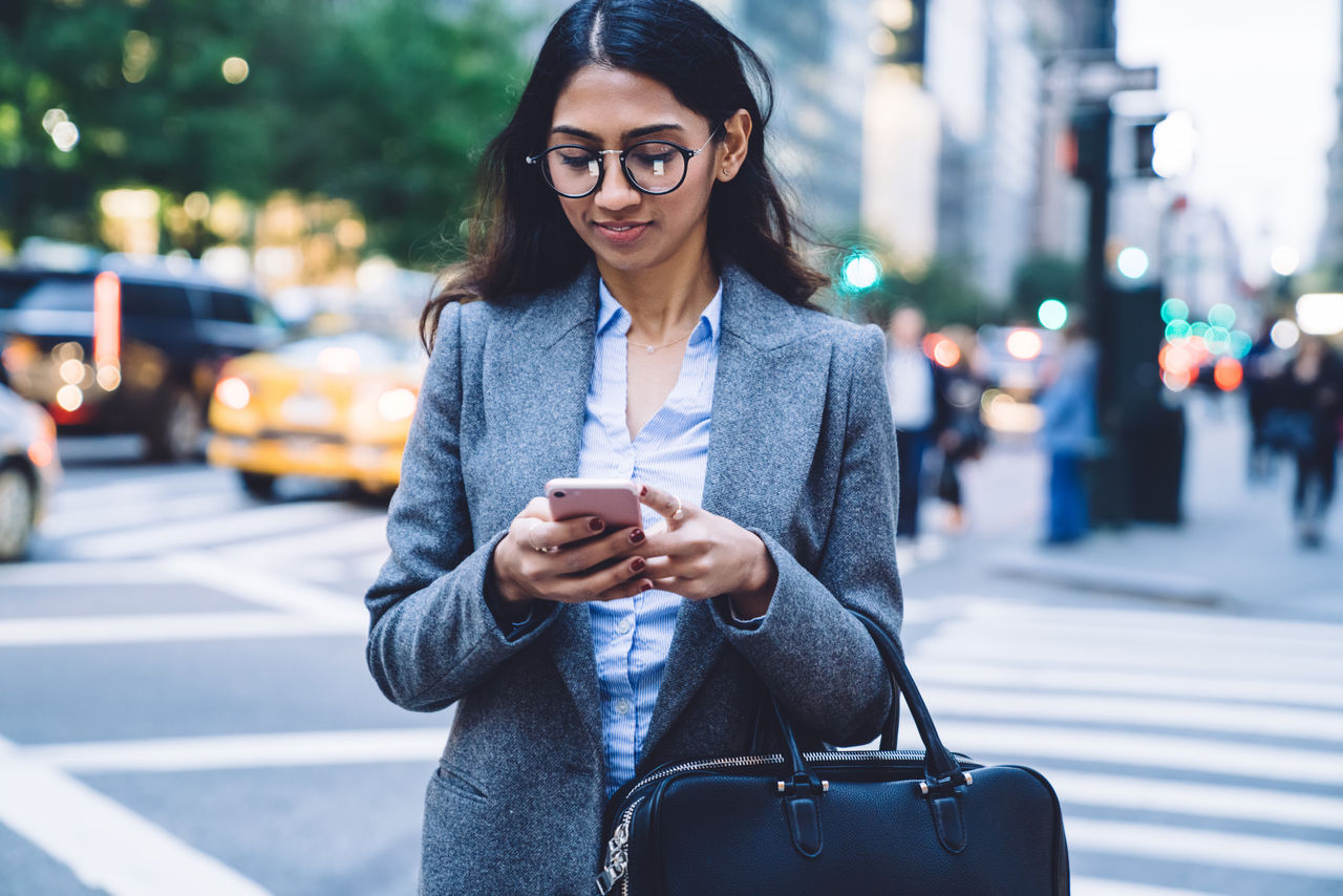 Young beautiful ethnic business woman with black hair and leather bag texting on mobile phone while crossing road in New York City